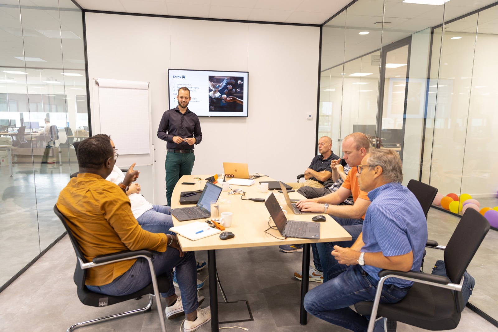 Trainer bij een scherm terwijl deelnemers aan een tafel met laptops meevolgen
