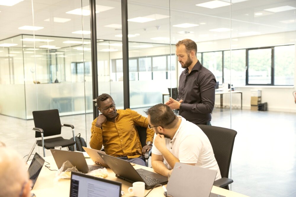 Collega's overleggen rond laptops aan een tafel in een kantoorruimte met glazen wanden.